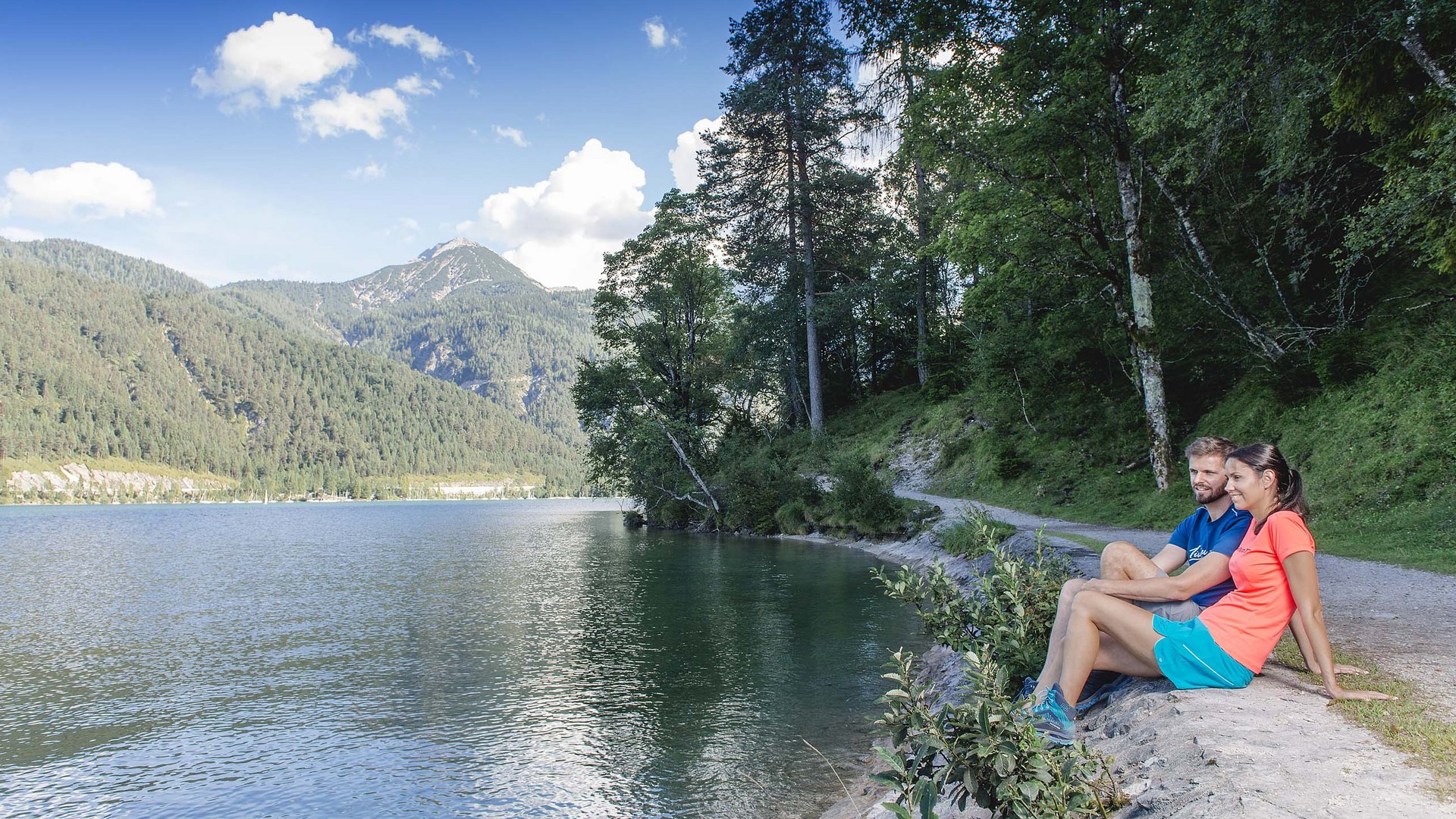Wandern am Achensee: traumhaft schön Paar sitzt am Seeufer mit Bergblick und Wald im Hintergrund