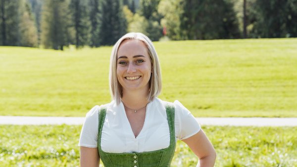 Die Karlwirt-Crew Frau in grünem Dirndl lächelnd vor grüner Wiese und Wald