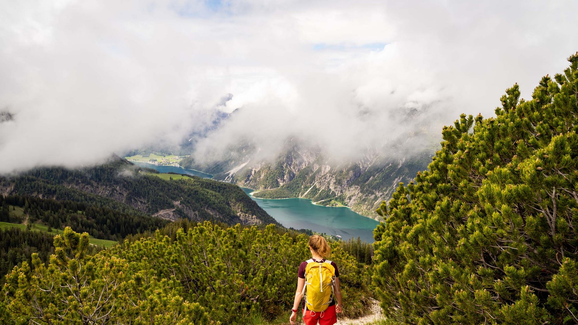 Lake Achensee: day trip options and more Hiker with yellow backpack on mountain trail overlooking forested lake and foggy mountains