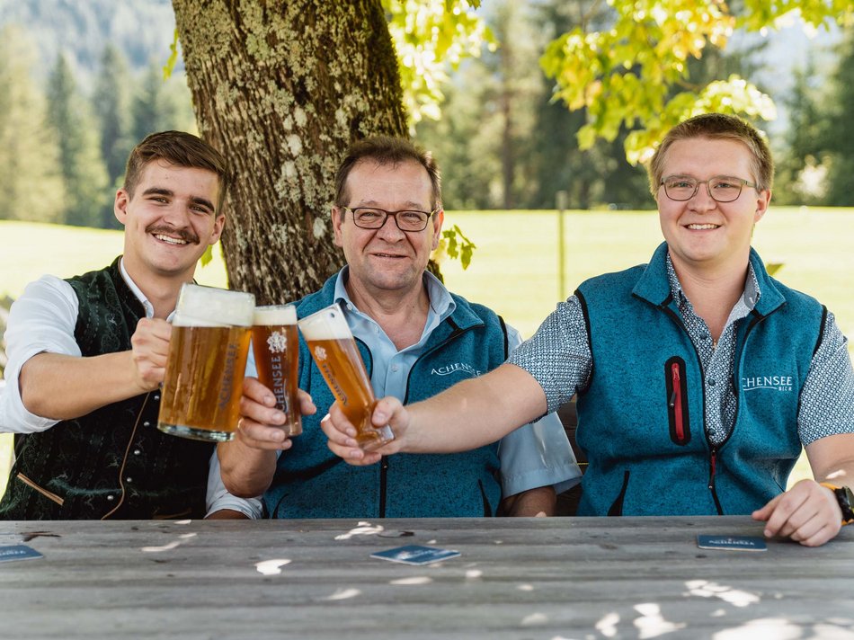 Das Achenseebier des Karlwirt Drei Männer stoßen mit Biergläsern im Freien unter einem Baum an