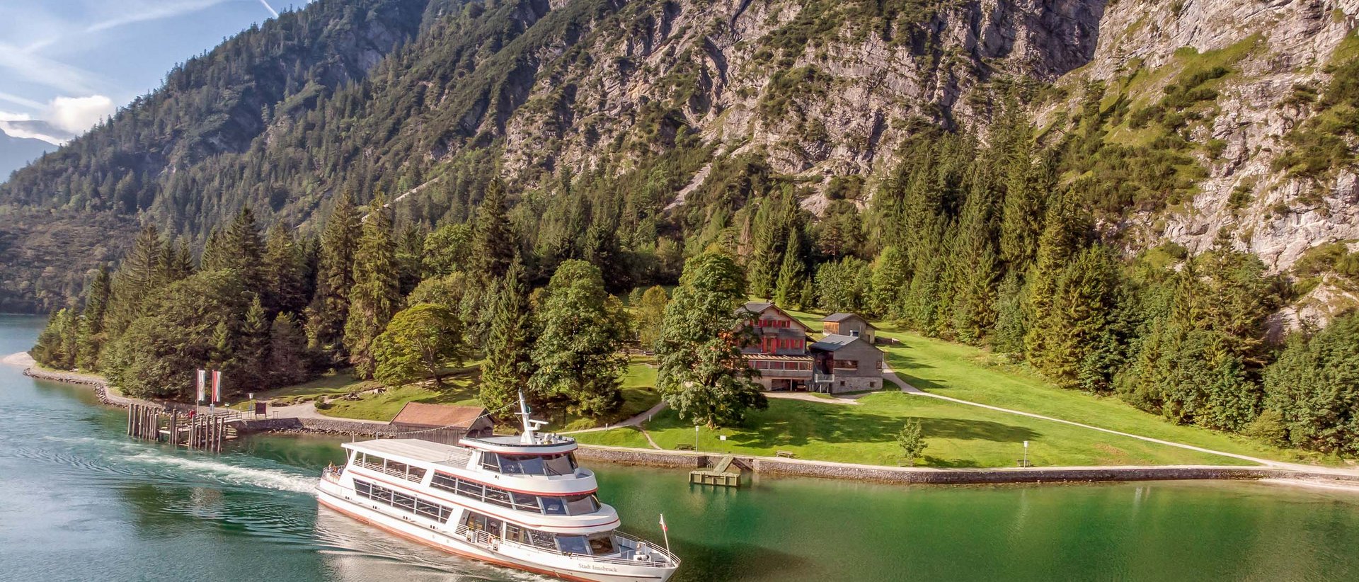 Entdeckt die versteckten Naturschätze Tirols. Personenschiff fährt über grünen See vor Alpenbergen und Wäldern
