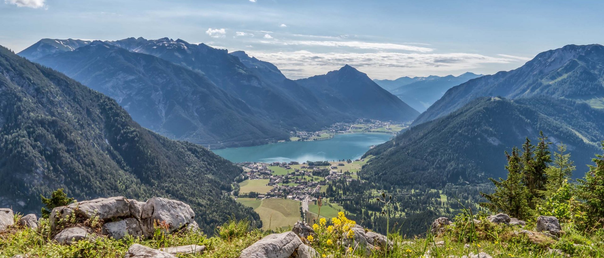 Sommerwanderung Blick auf Bergen, Tal und See mit Felsen und Blumen im Vordergrund