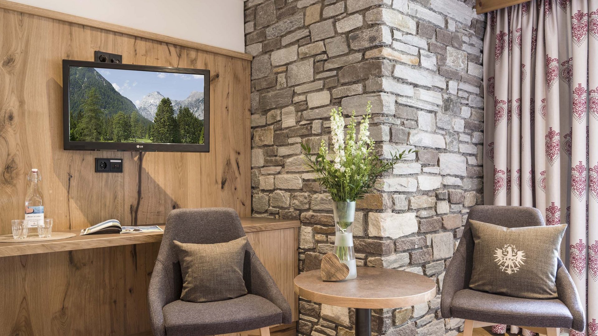 4-star hotel on Lake Achensee in Pertisau Cozy seating area with two chairs, round table, TV, and stone wall background