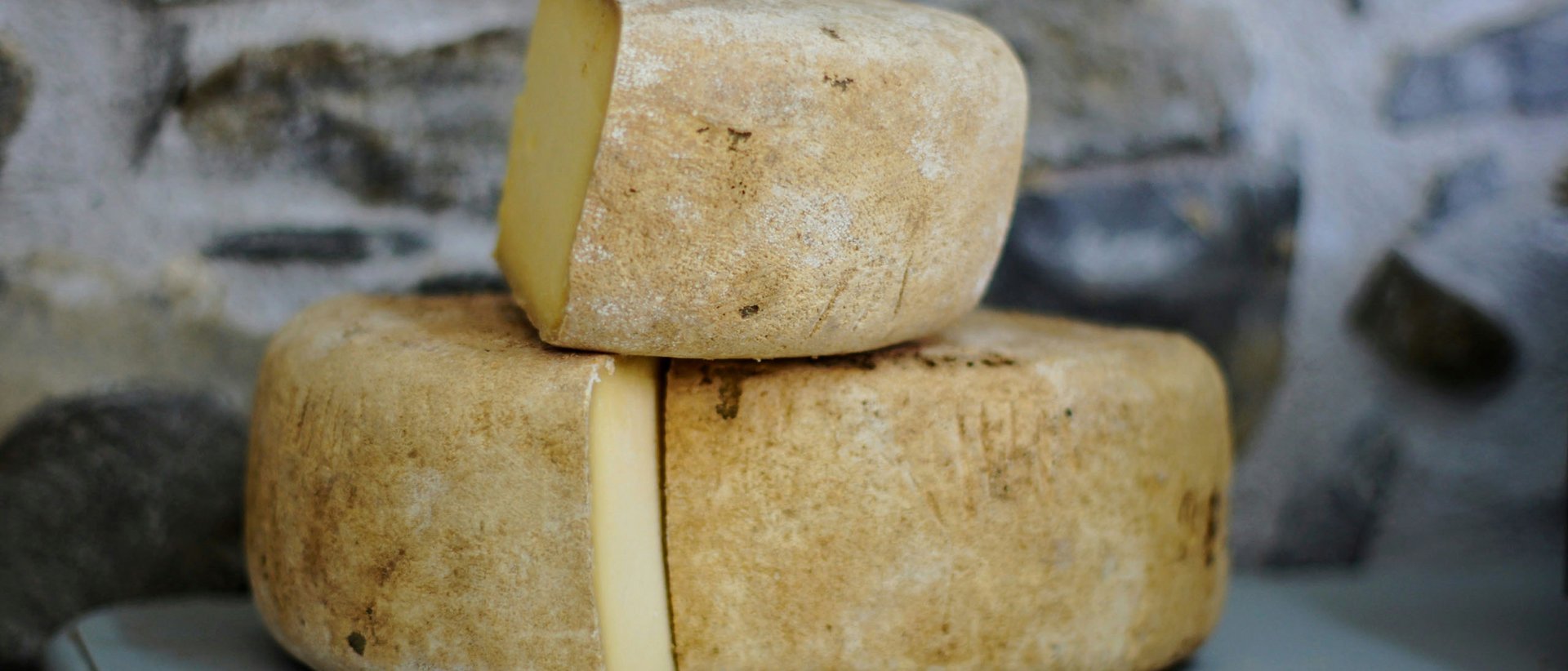 Deftig, herzhaft, lecker Drei Laibe gereifter Käse mit rötlicher Rinde auf einem Tisch vor Steinwand