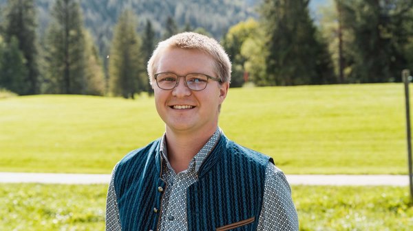 Die Karlwirt-Crew Junger Mann mit Brille und Trachtenweste in einer grünen Berglandschaft