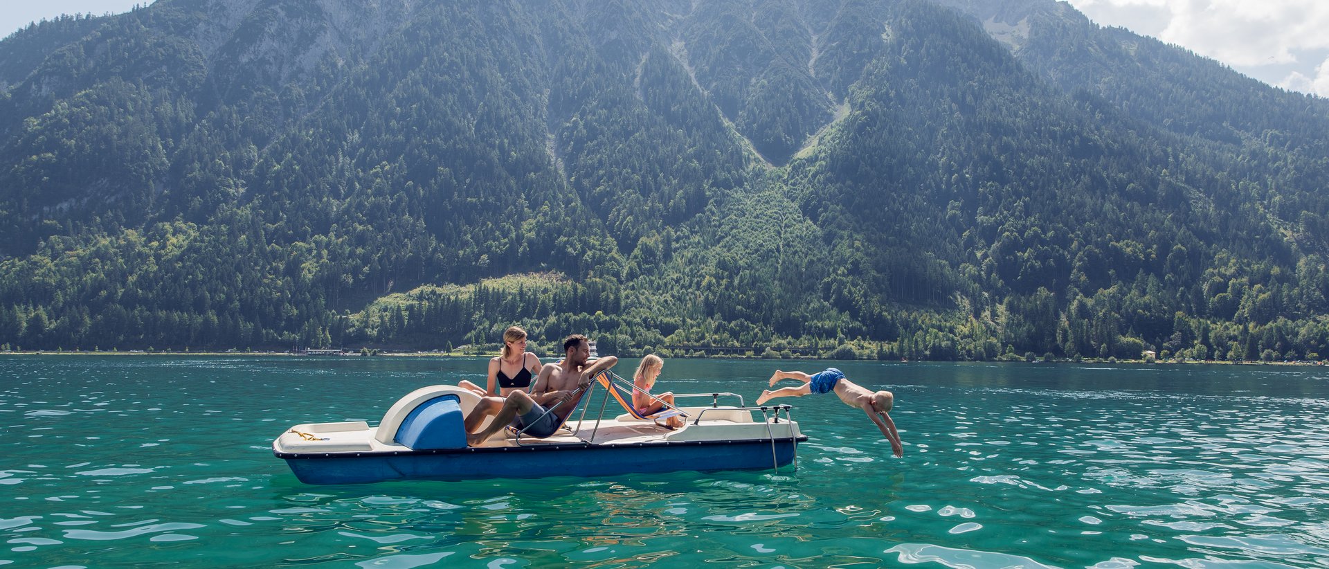 Tirols größter See wird im Sommer zum Spielplatz! Familie auf Tretboot vor Bergkulisse, Kind springt ins Wasser