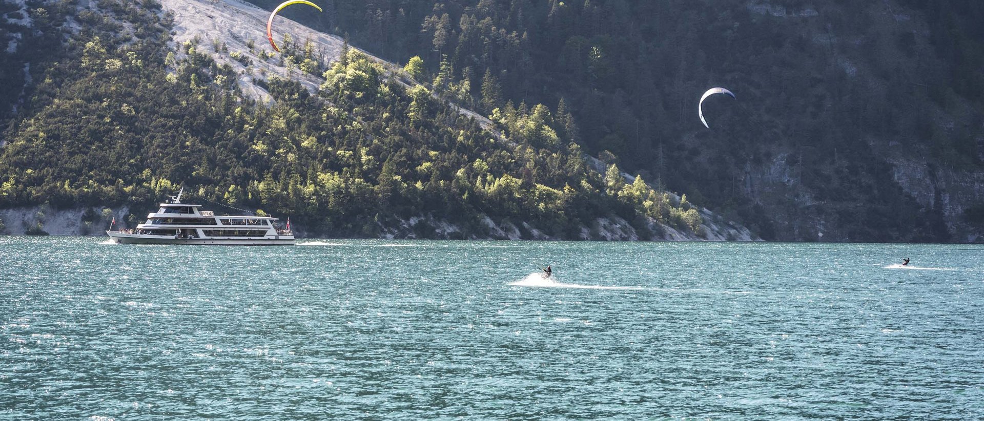 Abtauchen und genießen Boot und Kitesurfer auf See vor bewaldetem Berg
