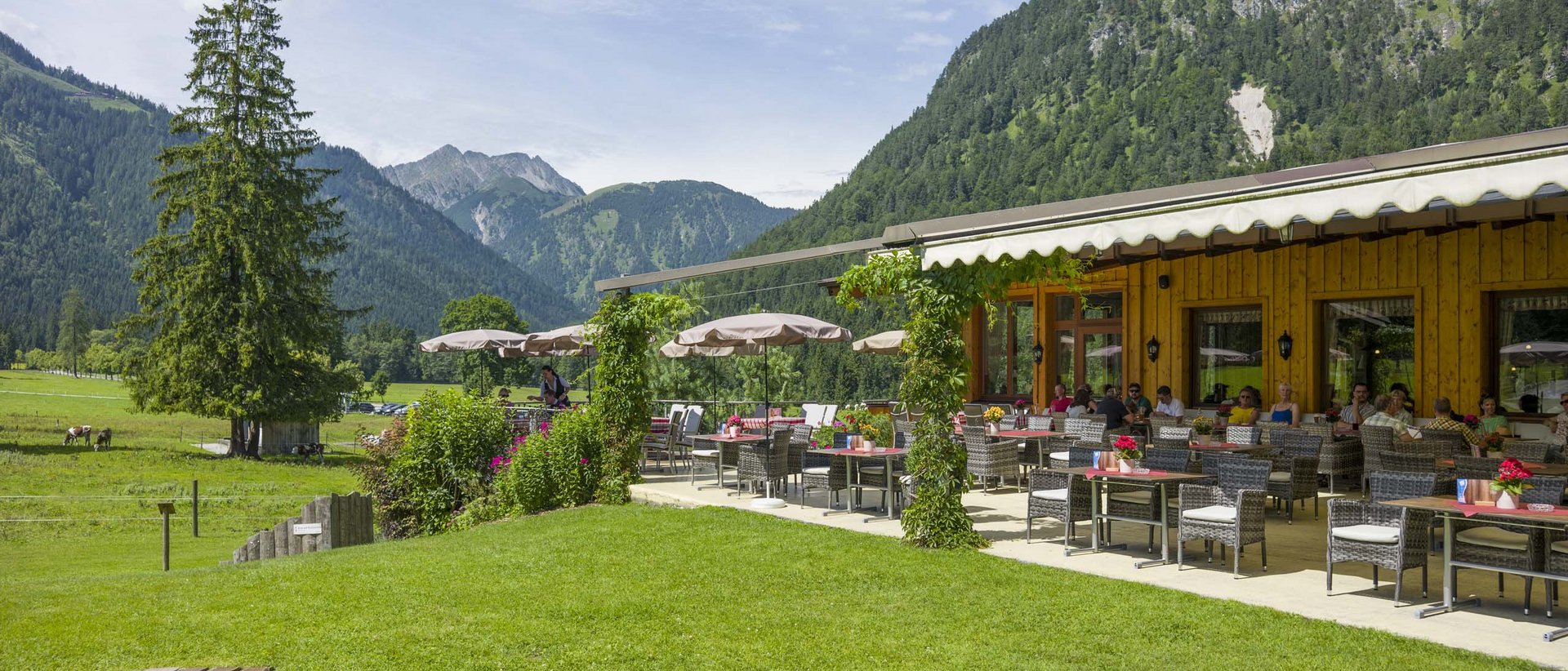 Einkehren und bleiben wollen Terrasse eines Berghotels mit Gästen, grüner Wiese und Bergen im Hintergrund