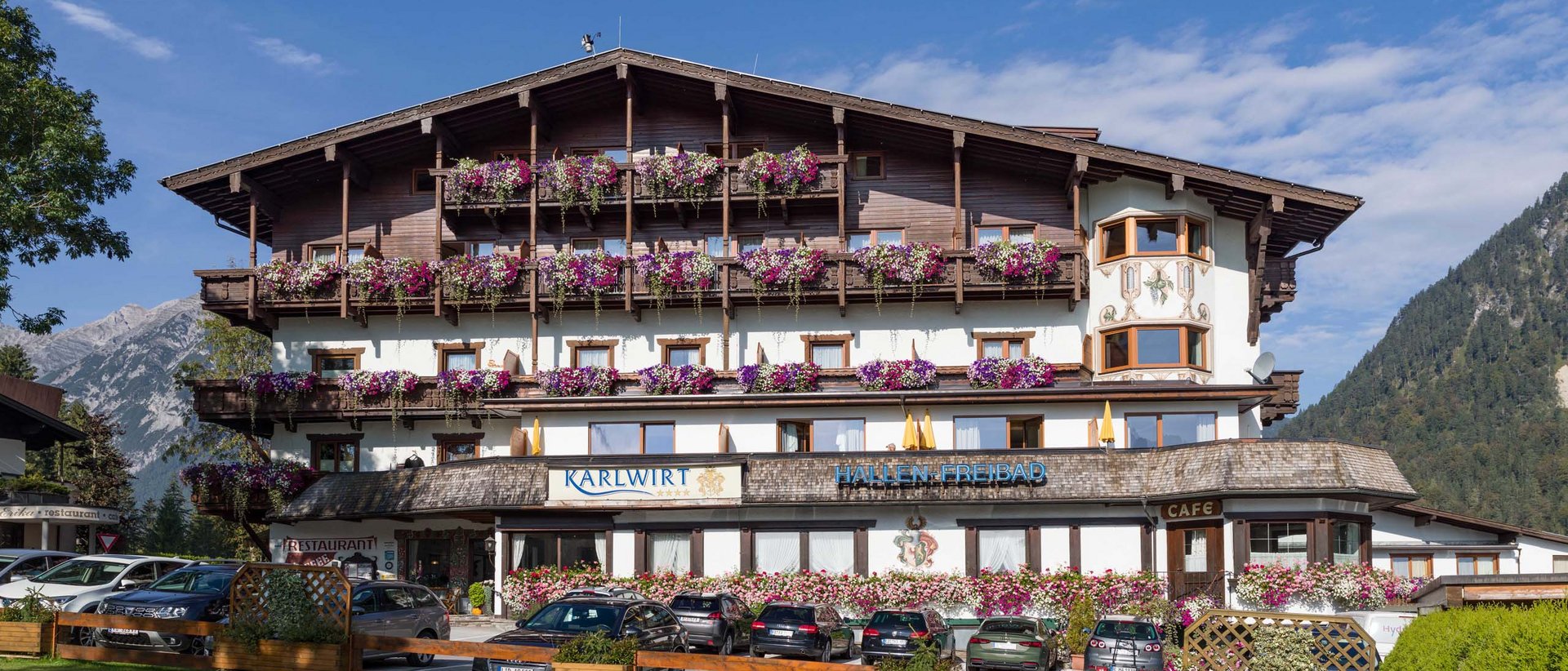 Neu im Karlwirt: unsere Website! Hotelgebäude mit Blumenschmuck und Autos vor dem Eingang in den Alpen