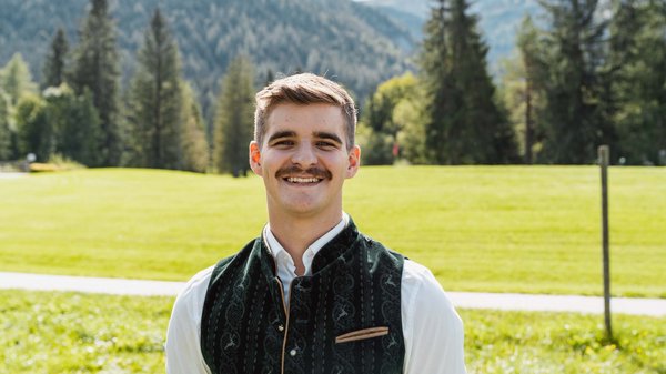 Die Karlwirt-Crew Junger Mann mit traditioneller Tracht vor Berglandschaft und grüner Wiese