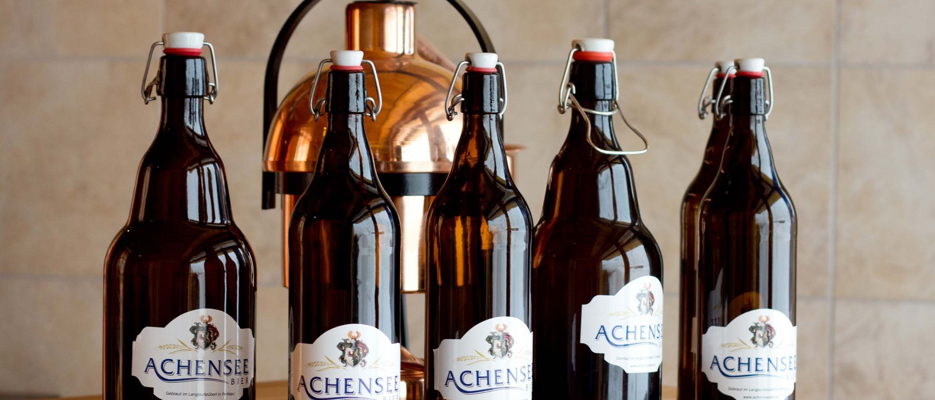 Die beliebteste Kleinbrauerei Tirols Sechs braune Bierflaschen Achensee vor Kupferbierkrug auf Holztisch